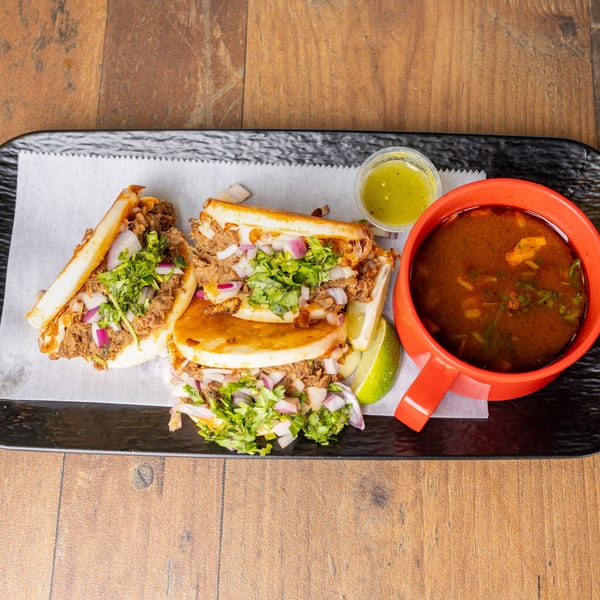 Three birria tacos with cilantro, onions, lime, green salsa, and a cup of consommé.