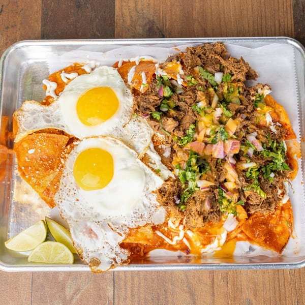 Chilaquiles with two fried eggs, shredded meat, onions, cilantro, and lime wedges on a metal tray.