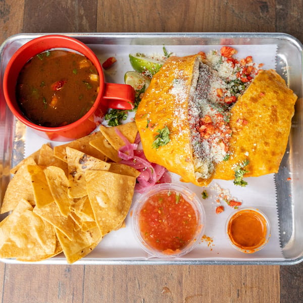 A metal tray with a large burrito, a red cup of soup, tortilla chips, and two small sauce cups.