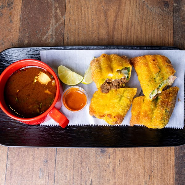 Birria egg rolls with consommé, lime wedges, and hot sauce on a black tray.